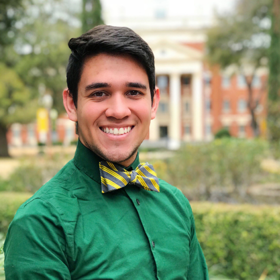 Man smiling with short brown hair, wearing a green button-up top with a striped yellow and grey bow-tie, standing outside in front of an administrative building and some greenery.